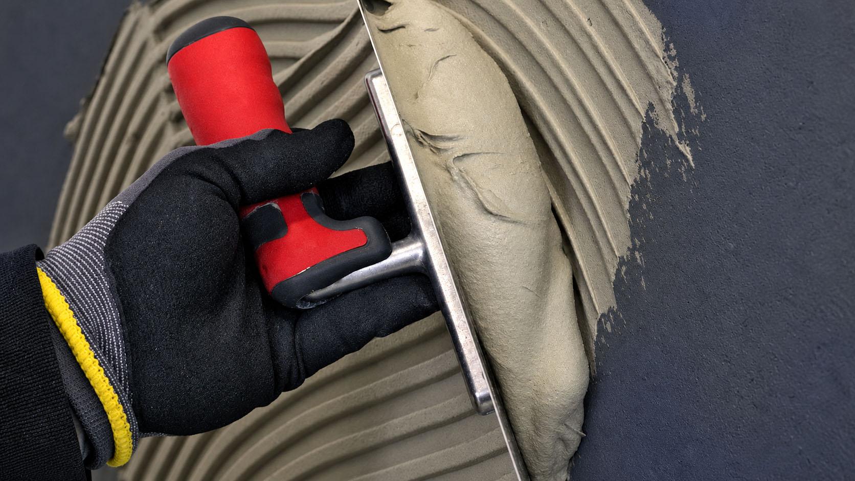 Hand mit Arbeitshandschuh trägt PCI Flexmörtel Light mit einer Zahnkelle auf einen dunklen Untergrund auf.